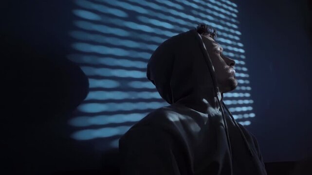 A young man wearing a hoodie sits in a dark room at night looking upward in deep thought or concern. Depression and burnout concept. Space for text