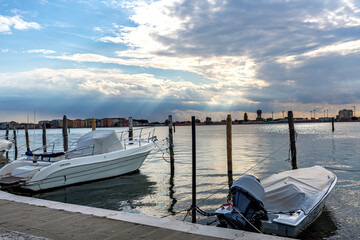 Fishing boats are lined up along a waterfront promenade, in Sottomarina, Italy,  displaying colorful details and textures, with a serene atmosphere and clear blue sky enhancing the scene