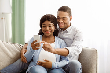 Loving black expecting parents shopping online from home, using mobile application on smartphone,...