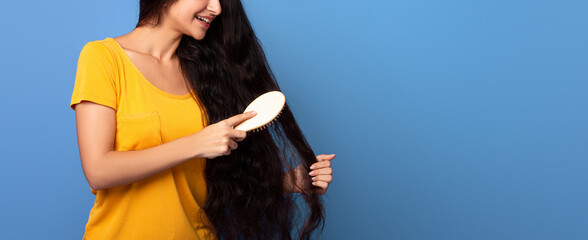 Portrait of smiling young indian woman looking at her long beautiful curly and wavy dark hair, combing with wooden brush, standing isolated on purple studio background, free copy space