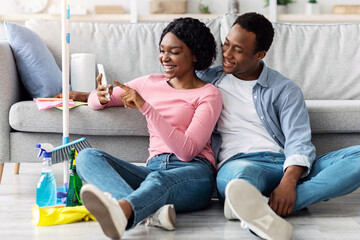Cheerful black couple with cleaning tools sitting on floor in living room, using smartphone,...