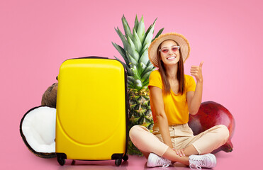 A young woman sits with a thumbs-up next to a bright yellow suitcase. Behind her are tropical...
