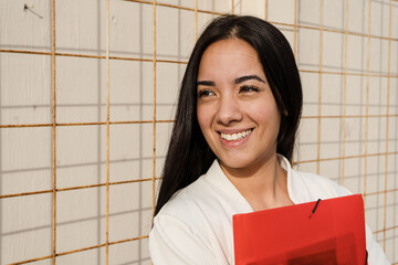 Young professional woman smiling and holding a red folder near a metallic grid