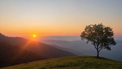 Solitary tree stands on green hill against warm sunrise sky. Layers of mountains recede into distance. Dawn light breaks over landscape, creating tranquil and peaceful atmosphere for nature lovers.