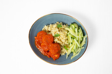 Bowl with bulgur, meatballs in tomato sauce, and fresh cucumber sticks on a white background. Top view