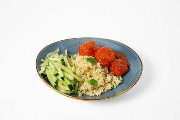 Bowl with bulgur, meatballs in tomato sauce, and fresh cucumber sticks on a white background. Top view