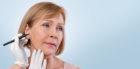Plakat Doctor drawing marks on senior woman's face for cosmetic surgery operation on light studio background. Mature lady getting ready for aestetic operation or face lifting