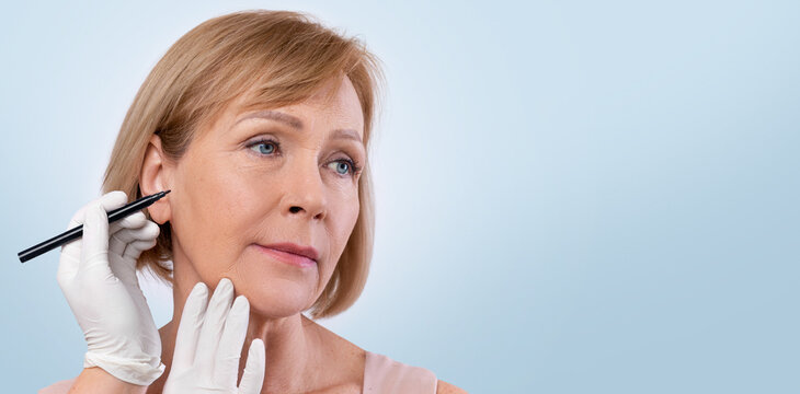 Fototapeta Doctor drawing marks on senior woman's face for cosmetic surgery operation on light studio background. Mature lady getting ready for aestetic operation or face lifting