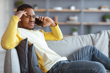 Upset black guy sitting on couch, having conversation on mobile phone, looking at copy space. Angry african american young man talking on smartphone, fighting with girlfriend