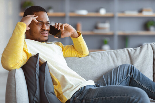 Upset black guy sitting on couch, having conversation on mobile phone, looking at copy space. Angry african american young man talking on smartphone, fighting with girlfriend - Powered by Adobe