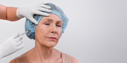 Mature lady in medical disposable hat receiving beauty shot from cosmetologist on light studio...