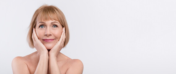 Beautiful half-naked elderly woman touching her face, applying skin care product on white studio...