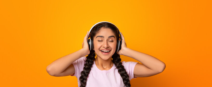 Good Sound. Portrait of excited indian woman wearing and touching wireless headphones, listening to music with closed eyes, female enjoying favorite song, isolated over yellow studio background