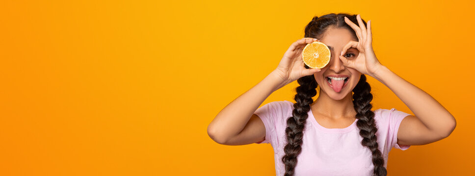 Portrait of joyful indian woman covering one eye with half of citrus orange fruit, looking at camera through fingers, doing okay gesture, imitating binoculars, isolated over yellow studio background