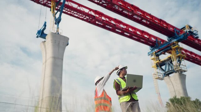 Two men are standing on a construction site, one of them holding a laptop. They are looking at the bridge they are working on