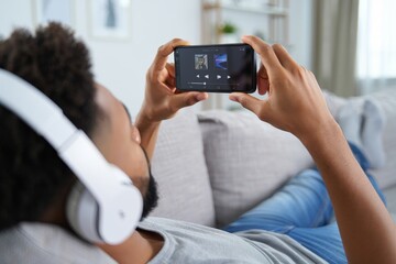 Man listening to music on phone with headphones relaxing on sofa at home streaming app