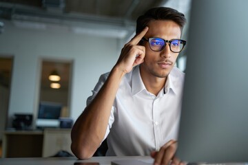 Man working late at night on computer wearing glasses thinking hard at the office