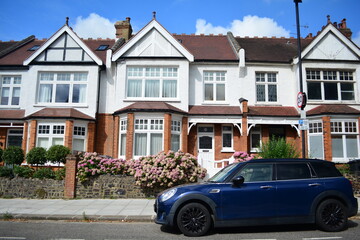 Beautiful terraced houses line the street under a bright blue sky. A black car is parked along the curb, and colorful flowers create a lively atmosphere around the homes