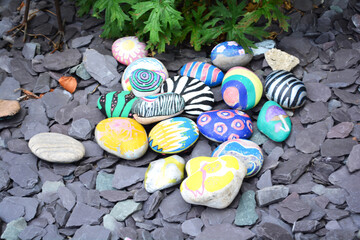 Colorful painted stones display intricate designs and patterns scattered across dark pebbles in a sunny garden. Vibrant hues and unique artwork make this a creative outdoor decoration