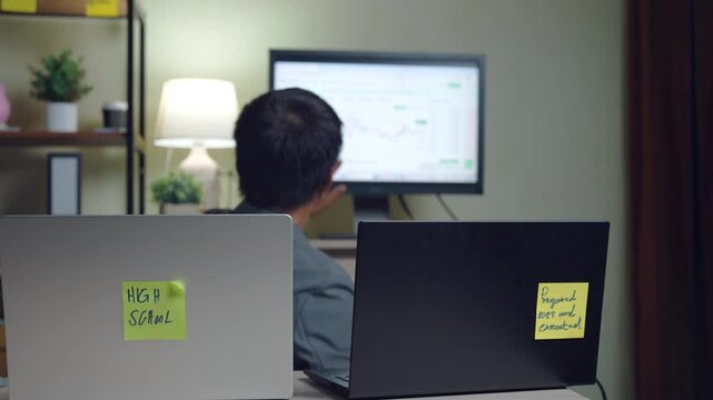 A man is sitting at a desk with two laptops. One laptop has a sticky note on it that says "SSG"