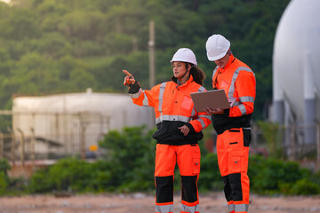 Two engineers in safety gear review project plans on a laptop and use a walkie-talkie at an...