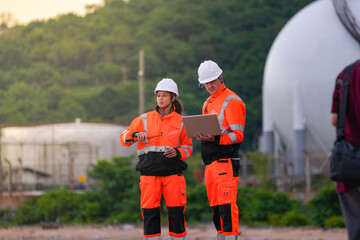 Two engineers in safety gear review project plans on a laptop and use a walkie-talkie at an...