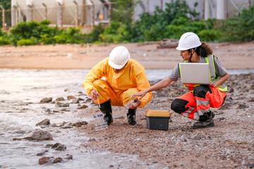 Two environmental engineers in safety gear work at a shoreline near gas storage tanks. One collects water samples, while the other records data on a laptop during site inspection.