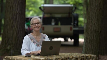 Portrait of smiling mature woman using a computer and phone at a campground, enjoying the outdoors...