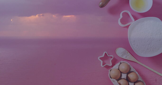 Fototapeta Displaying sea under purple-pink sky and baking setup on pink surface with flour bowl, copy space