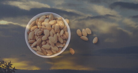 Reflecting white ceramic bowl holding almonds on reflective glass table at dusk, showing cloudy sky