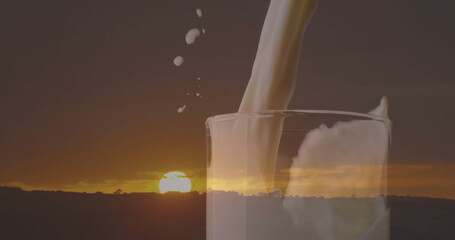 Pouring milk into transparent glass at rural dusk, sun refracting droplets on hill silhouettes