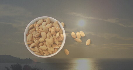 Overflowing white ceramic bowl sitting on shoreline at seaside sunset, with floating whole almonds