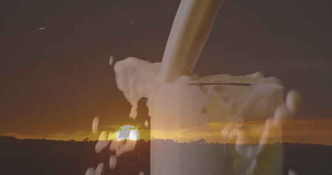 Pouring milk stream into clear glass, frothing overflow on rural horizon at sunset with tree line