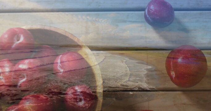 Presenting wooden bowl holding red plums on weathered kitchen table, with sandy shoreline overlay
