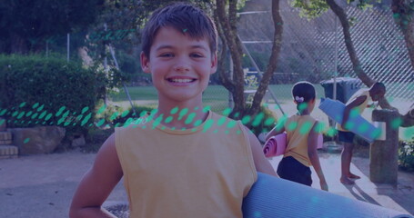 Smiling boy carrying blue yoga mat at outdoor playground, with water fountain and stone bench