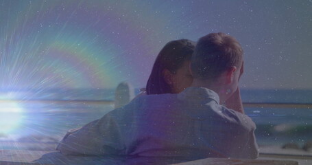 Hugging couple leaning toward ocean on boardwalk, with wooden bench and railing beneath sunflare