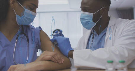 Injecting doctor administering vaccine into nurse in scrubs' arm at clinic, with syringe and vials