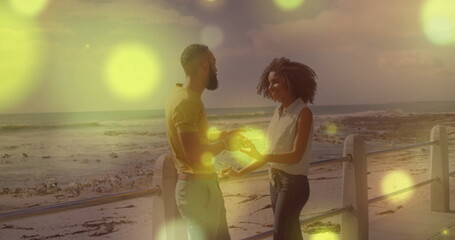 Standing couple holding hands on seaside promenade at sunset, with metal railing and ocean waves