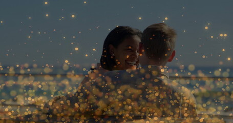 Smiling woman hugging man on waterfront deck, with metal railing and golden bokeh lights