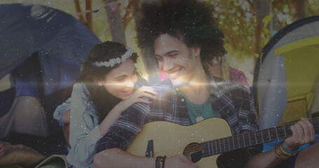 Playing man strumming acoustic guitar at forest campsite with dome tents floral headband bracelets