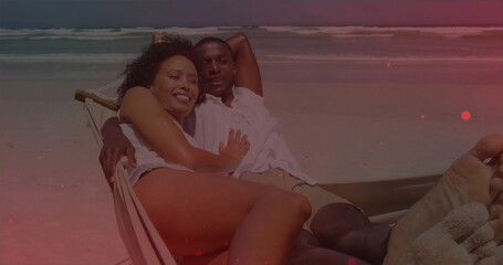 Lying couple wearing summer clothing on hammock lounger at sandy beach, ocean waves rolling