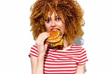 Young woman with curly hair eating a cheeseburger, showcasing a joyful expression in a vibrant, colorful outfit on a bright white background