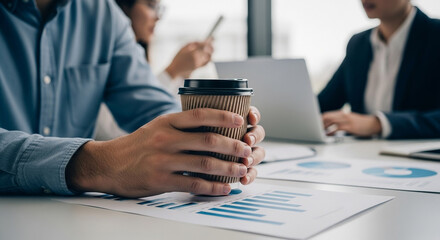 A person holding a coffee cup while colleagues work on a project in an office setting.