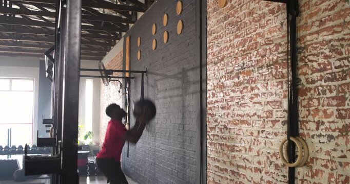 African American man entering gym picking up black med ball, squatting and throwing at wall target