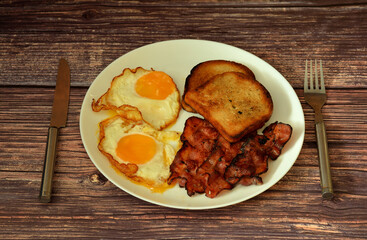 A plate of nutritious breakfast, two fried eggs, bacon and toast on a wooden table.