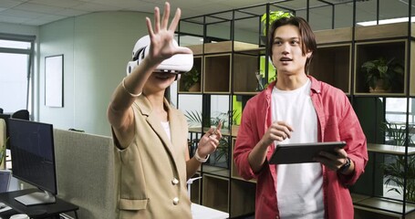 Upon donning VR headset, Asian coworkers gesturing in office using tablet for VR demonstration
