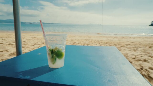 Close-up of a person's hand grabbing a refreshing Brazilian caipirinha cocktail with lime and ice, served on a beach table with ocean and sky backdrop during sunny summertime relaxation.