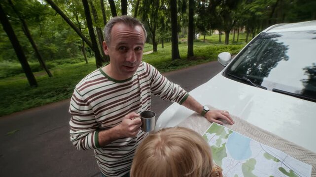 Tilt up and down of active family of father, mother and son standing by car hook, having tea and discussing their location and further route on paper map
