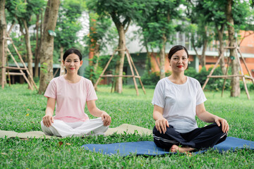 Two women sit in lotus position practicing yoga outdoors in a green park with trees in the background