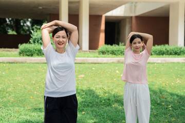 Two women in athletic wear stretching in a park with grass and buildings in the background